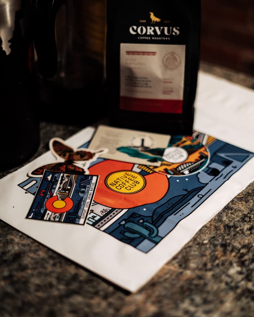 Specialty coffee from Corvus sitting on a shipping bag from Native Coffee Club along with stickers and free drink cards on a counter