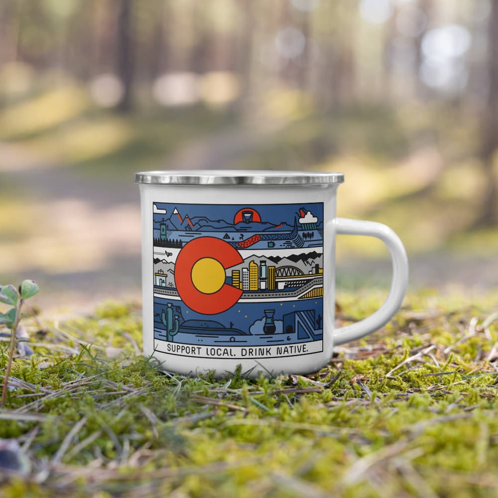 White Enamel camping mug with the Colorado flag, scenery, and specialty coffee themes intertwined displayed on moss in the woods. At the bottom of the mug it says Support Local. Drink Native. 
